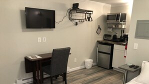 A compact hotel room with a bed, a desk with a chair, a television, a kitchenette, and a small table with a remote control.