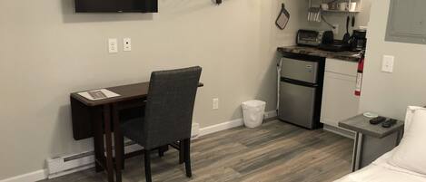 A compact hotel room with a bed, a desk with a chair, a television, a kitchenette, and a small table with a remote control.