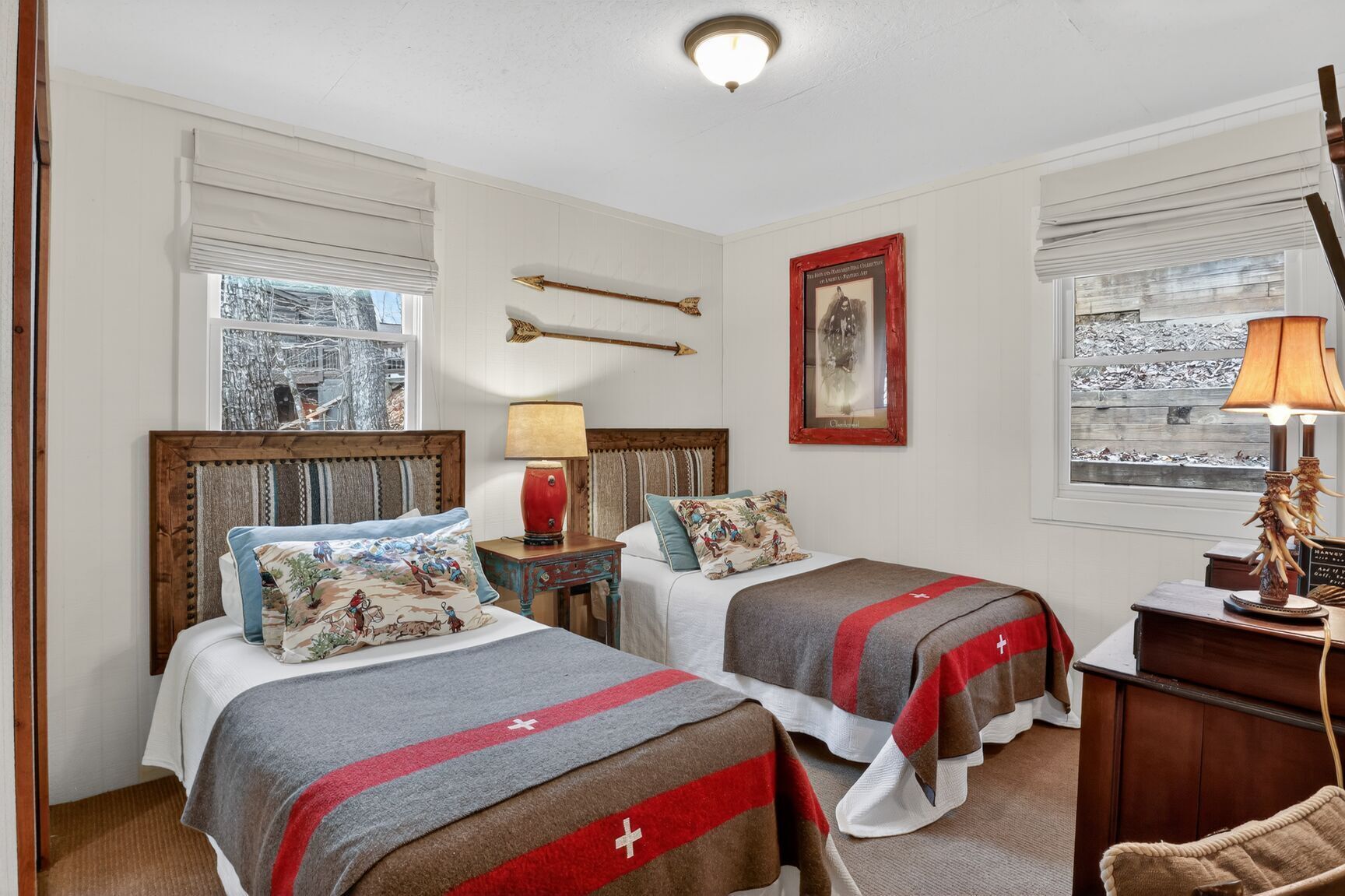 Una habitació d'hotel amb dos llits, un panell de capçalera de fusta, una tauleta de nit amb una làmpada, un quadre a la paret i una finestra amb vista a arbres.