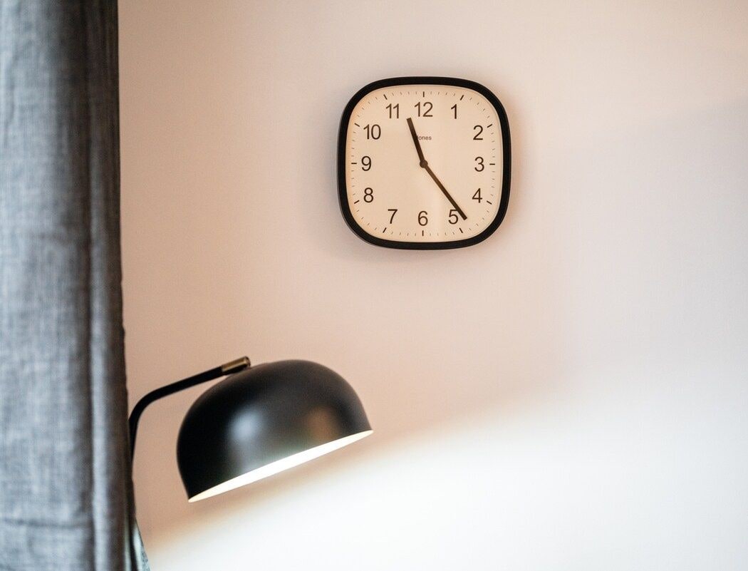 A wall-mounted clock with a minimalist design, a black desk lamp, and a logo in the bottom right corner.