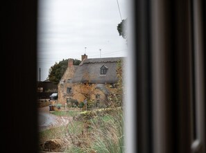 A traditional stone house with a thatched roof, visible through a window with a dark frame. - 3x Bedroom House (Sleeps 7x) (Oxfordshire)