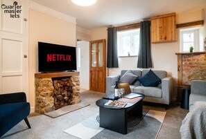 A cozy living room with a stone fireplace, a grey sofa, a coffee table with a magazine, and a TV displaying 'NETFLIX'. - 3x Bedroom House (Sleeps 7x) (Oxfordshire)
