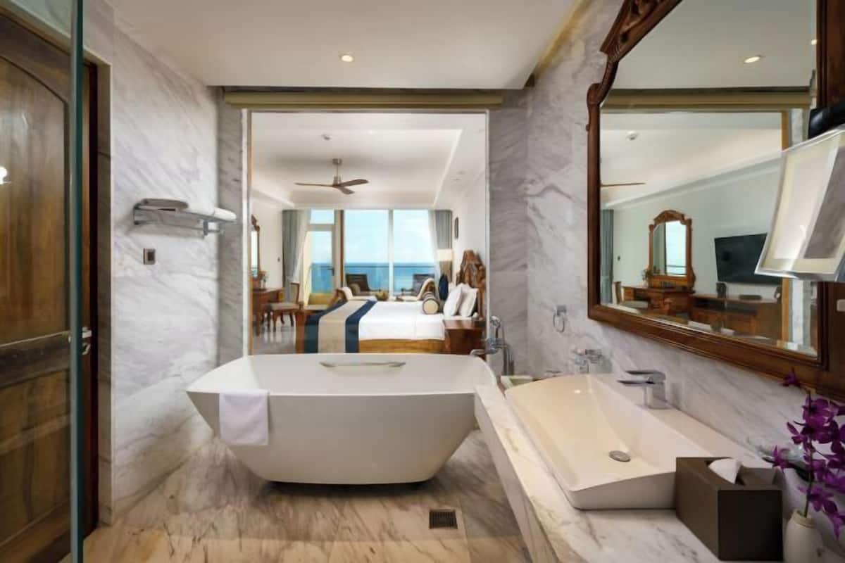 Une salle de bain moderne avec une baignoire sur pieds, un grand miroir et une vue sur une chambre à coucher avec un lit et une fenêtre donnant sur la mer.