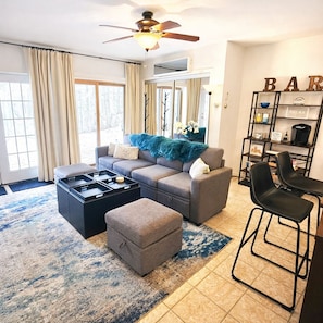A living room with a sofa, ottoman, coffee table, and a bookshelf with items on it.