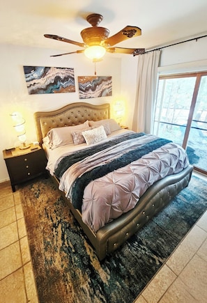 Une chambre Ă coucher avec un lit, un ventilateur de plafond, des Ćuvres dâart accrochĂ©es au mur et une vue par la fenĂȘtre.