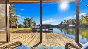 Um deck de madeira com cadeiras de vime, um compartimento envidraçado e vista para um lago com docas e um gazebo.