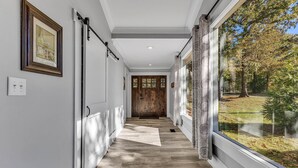Um corredor com piso de madeira, uma janela grande com vista para árvores e uma porta com painel de vidro.