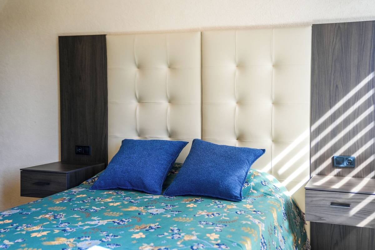 Une chambre à coucher avec une tête de lit capitonnée, une couvre-lit à motifs floraux, deux coussins bleus, une table de chevet et une armoire en bois.