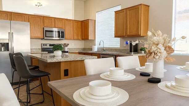 A modern kitchen with wooden cabinets, stainless steel appliances, and a dining area with white tableware and chairs. - Near stadiums and sports complex, Peoria Oasis (Peoria)