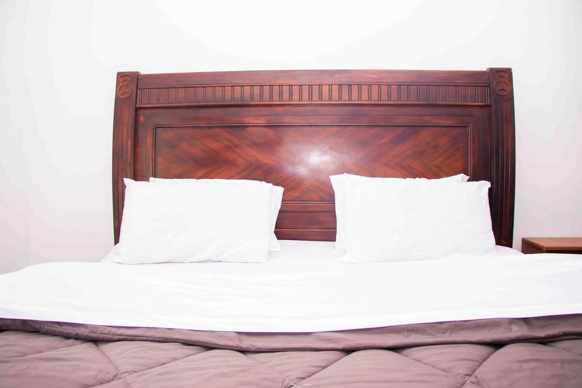 Een netjes opgemaakt bed met een houten hoofdbord, wit beddengoed en een klein nachtkastje ernaast.