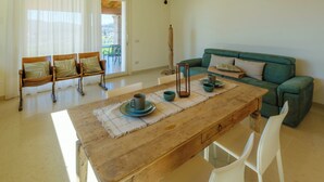 Een ruime kamer met een houten eettafel, witte stoelen, een groene bank en een groot raam met dunne gordijnen.