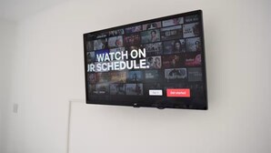 Una TV a schermo piatto montata su un muro bianco mostra un'interfaccia di servizio di streaming con diverse opzioni di programmazione.