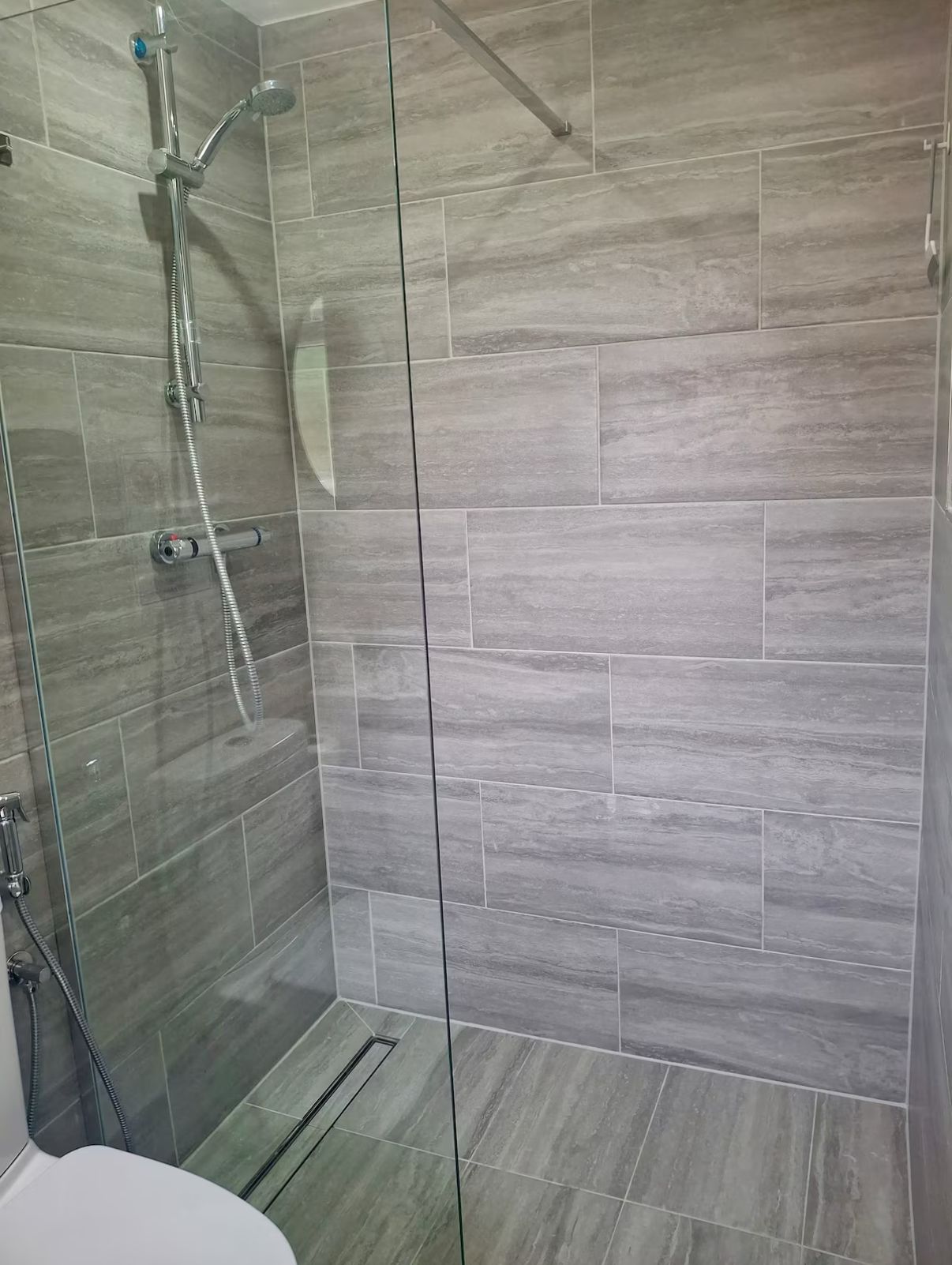 A modern bathroom with a glass shower enclosure, grey tiles, and a visible showerhead.