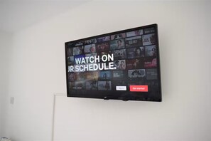 A flat-screen TV mounted on a white wall displaying a streaming service interface with various show options. - Cozy studio apartment near Heathrow (Northolt)
