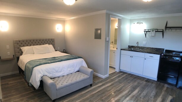 A bedroom with a bed, a bench, a kitchenette, and a bathroom visible through an open door.