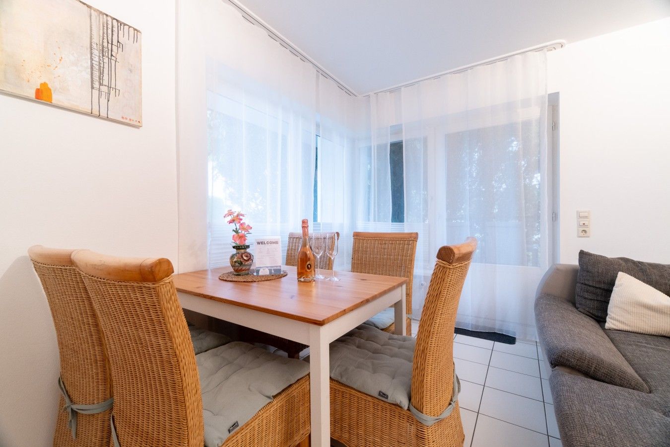 A modern living room with a dining area, featuring wicker furniture, a grey sofa, and a large window with sheer curtains.