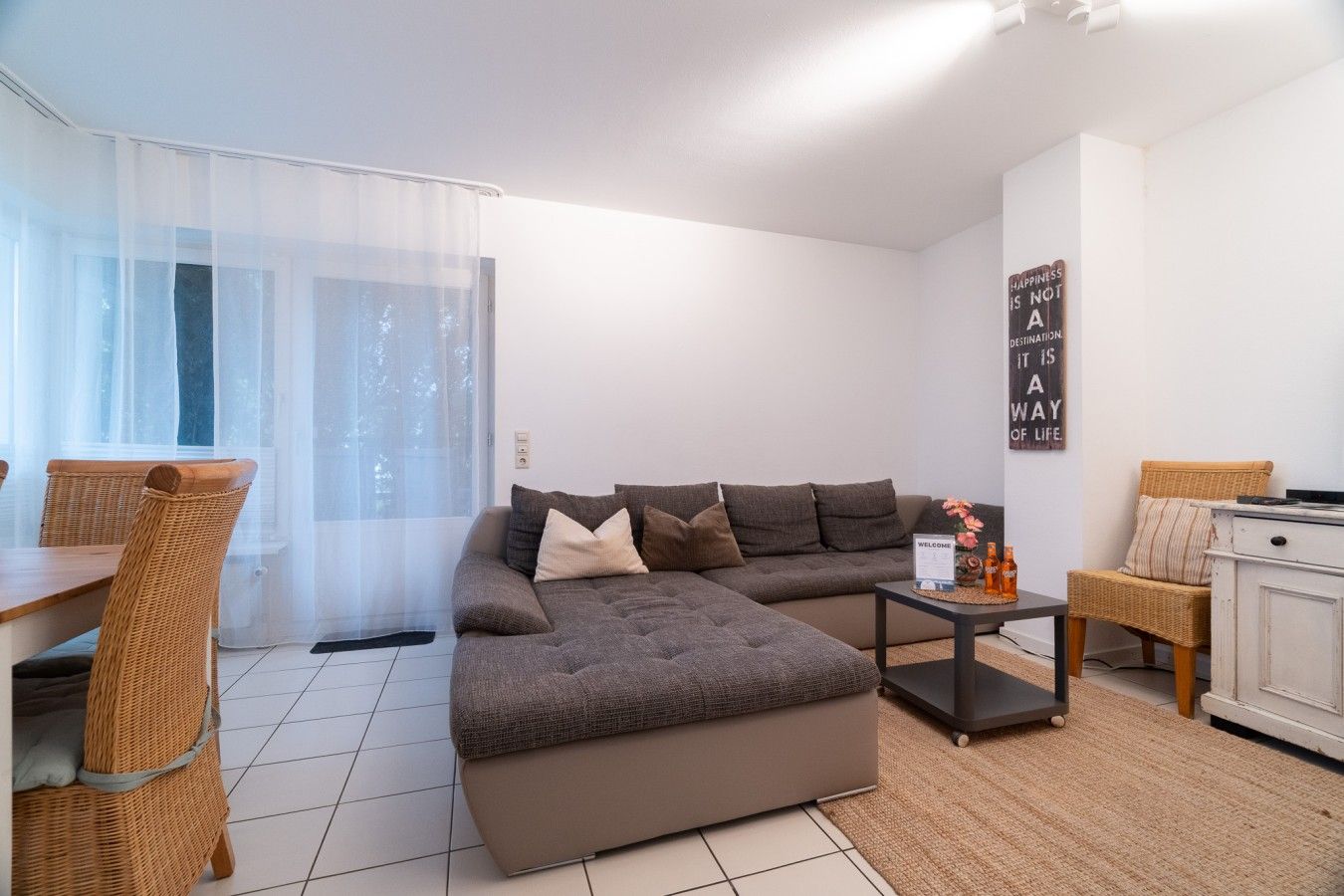 A modern living room with a sectional sofa, a dining table with chairs, and a small side table with a welcome sign.