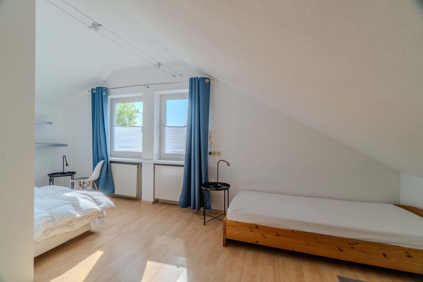 Ein kleiner, gut beleuchteter Raum mit schrägen Wänden, einem Bett, einem Schreibtisch, einem Stuhl und einem fenster mit Vorhängen.