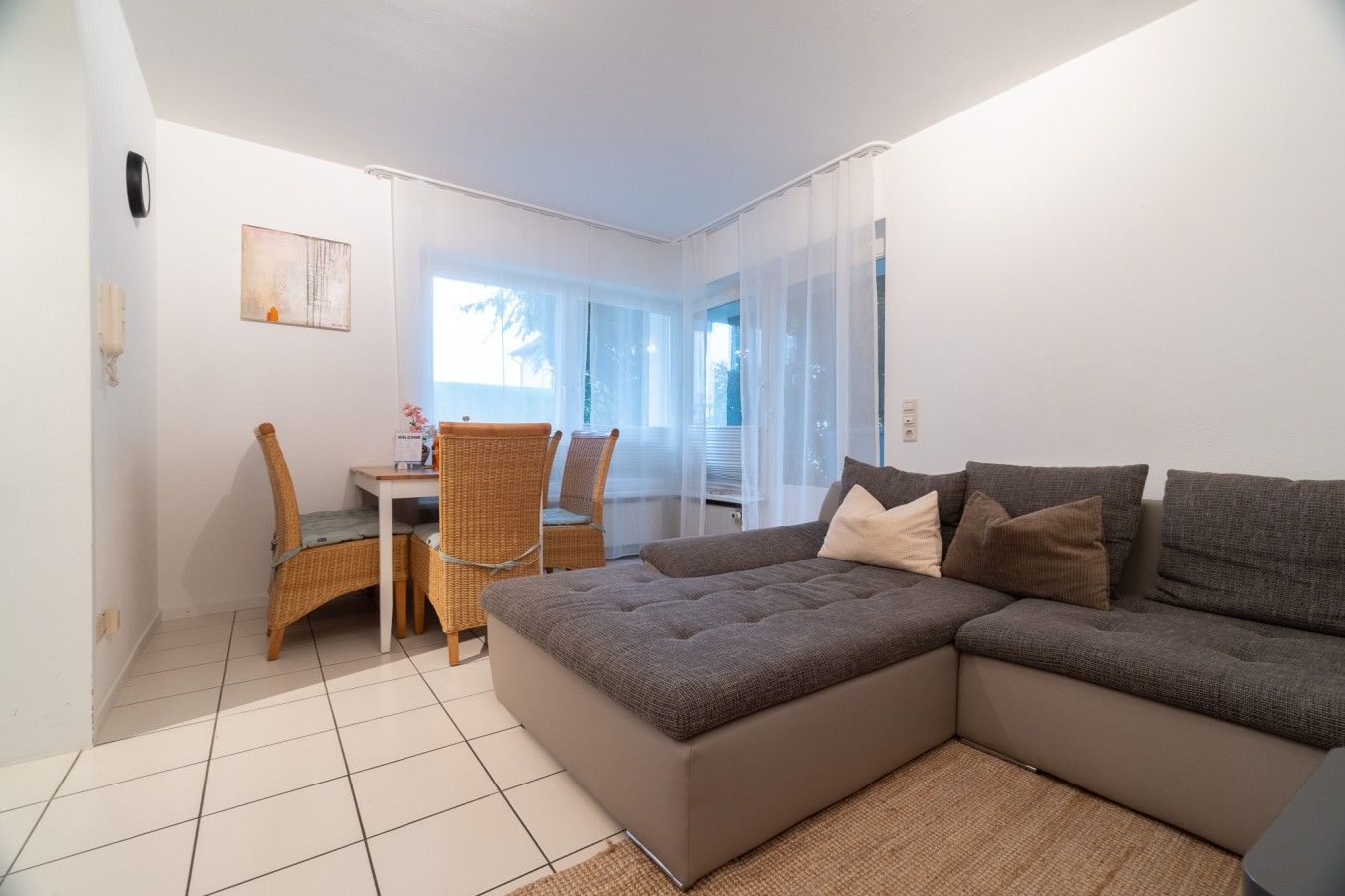 A modern living room with a sectional sofa, a dining area with wicker chairs, and a large window providing natural light.