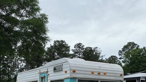 Une caravane ancienne, avec une porte et des fenêtres turquoise, garée sur un sol de gravier et entourée d’une clôture en bois peinte en blanc.
