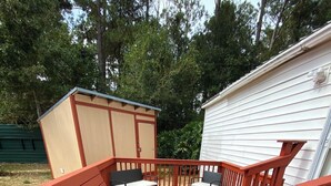 Eine rote Terrasse mit zwei Korbstühlen und einem kleinen Tisch, eine Gartenhütte und ein weißes Gebäude im Hintergrund.