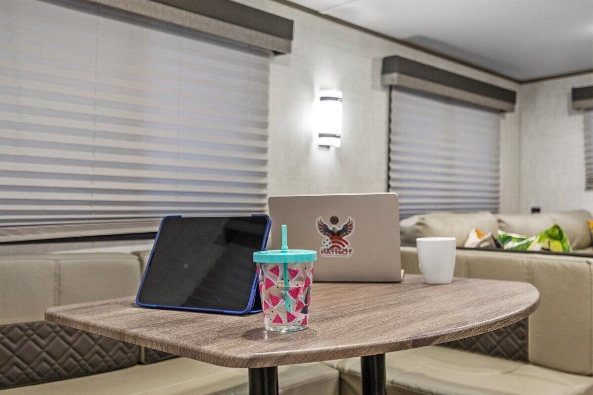 A laptop on a table with a drink and a cup in a room with a couch and blinds.