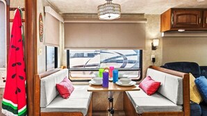 Interior compacto de autocaravana con kitchenette, dos zonas de estar con cojines, una mesa con vajilla y una ventana con vistas.