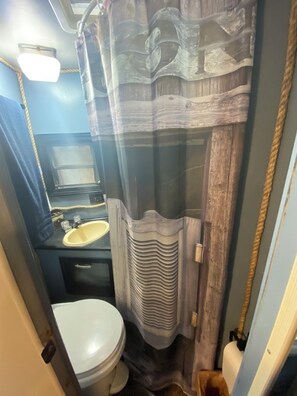 A compact bathroom with a toilet, sink, and shower curtain. - The "Bunkhouse" (western-themed glamper) sleeps 2 to 4 avg size people. There's even a bar with real western saddles for barstools! All new electric appliances, so super-easy to "glamp" in a beautiful, treed area of the park. Access to the park's am (Pace)