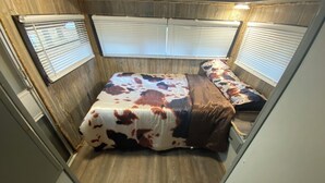 A compact RV bedroom with a bed, two windows, and a wooden floor.