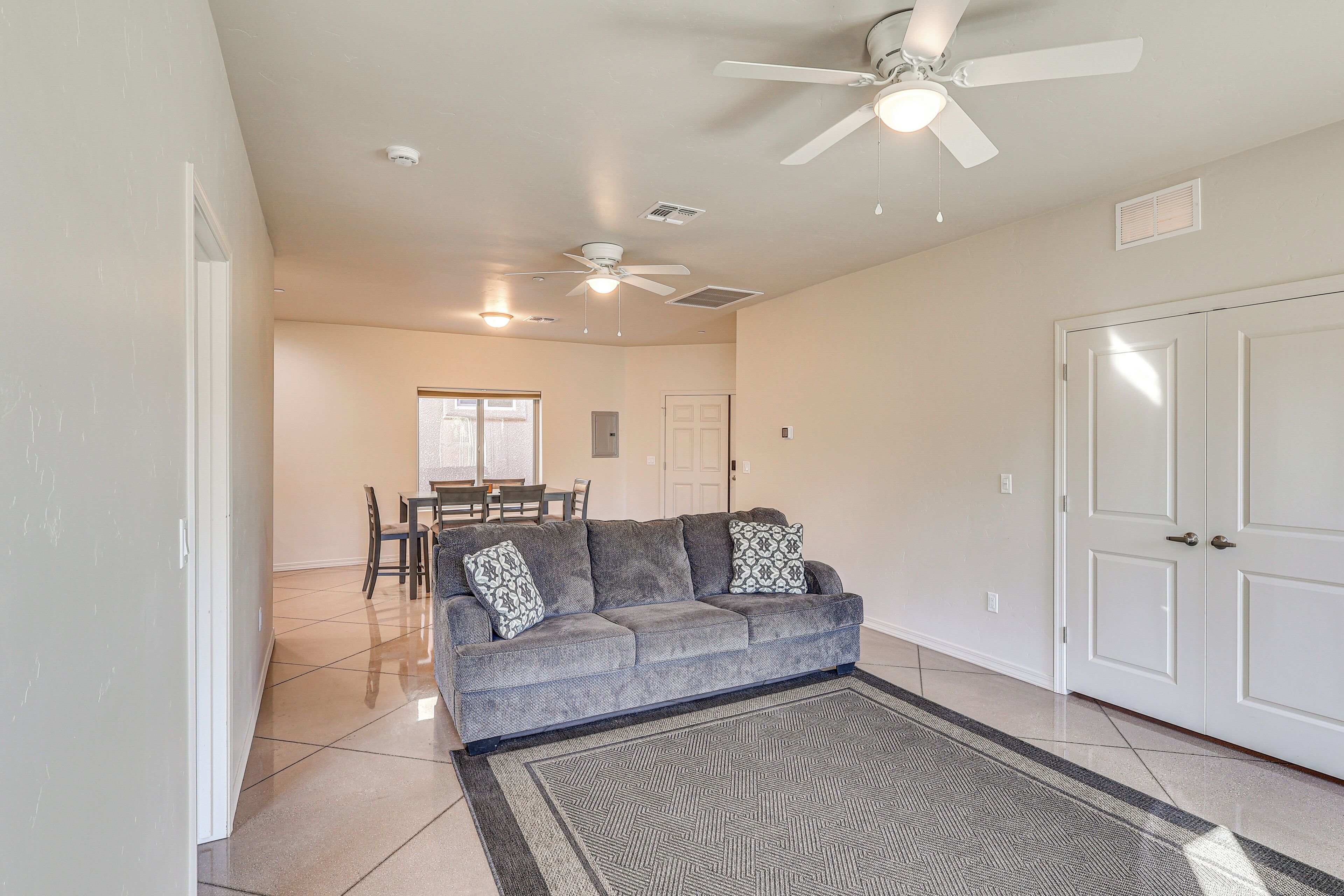 A living room with a gray sofa, patterned throw pillows, a patterned rug, a ceiling fan, and a dining area with a table and chairs.