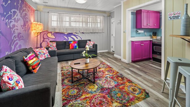 A modern living room with a gray sofa, colorful pillows, a wooden coffee table, and a vibrant rug. A kitchen with pink cabinets and a stove is visible in the background. - Modern & Bright Escape: Mins Beach, Pool, Hot Tub! (Sarasota)