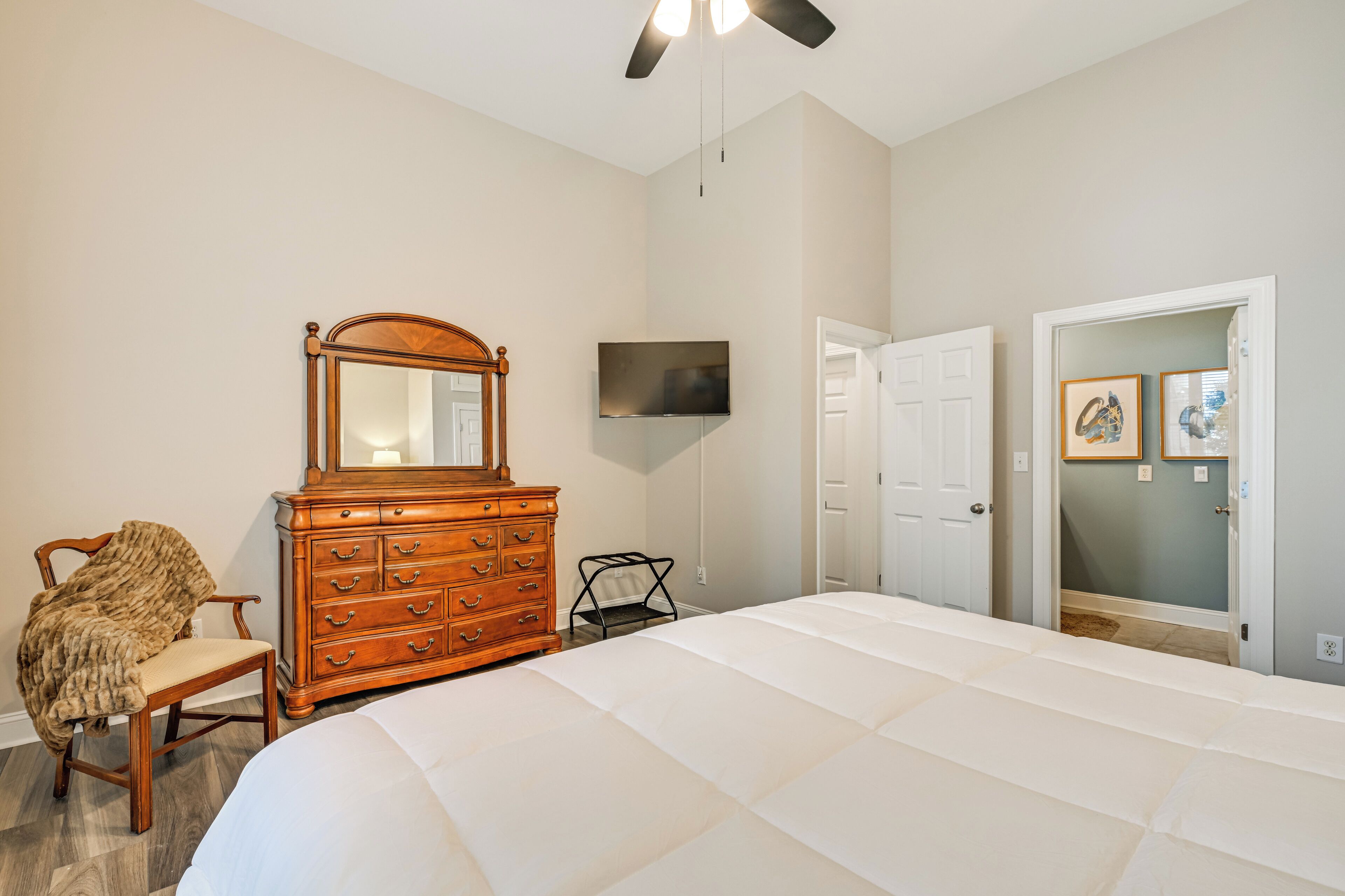 A bedroom with a large bed, a wooden dresser, a chair, a TV mounted on the wall, and a door leading to another room.