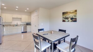 Una cocina moderna con armarios blancos, un refrigerador de acero inoxidable y un microondas. Hay una mesa de comedor con cuatro sillas y un cuadro en la pared.