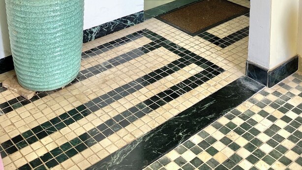 Un pasillo con suelo de baldosas, una maceta de cerámica grande y una puerta que da a un cuarto con una mesa y sillas de comedor.