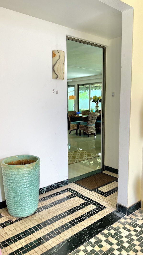 Un pasillo con suelo de baldosas, una maceta de cerámica grande y una puerta que da a un cuarto con una mesa y sillas de comedor.