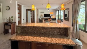 Una cocina con encimeras de granito, luces colgantes, una zona de comedor con sillas, un televisor y un ventanal con cortinas.