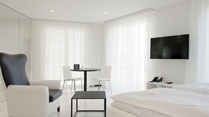 A modern hotel room with a large bed, a desk with a chair, a TV mounted on the wall, and large windows with sheer curtains.