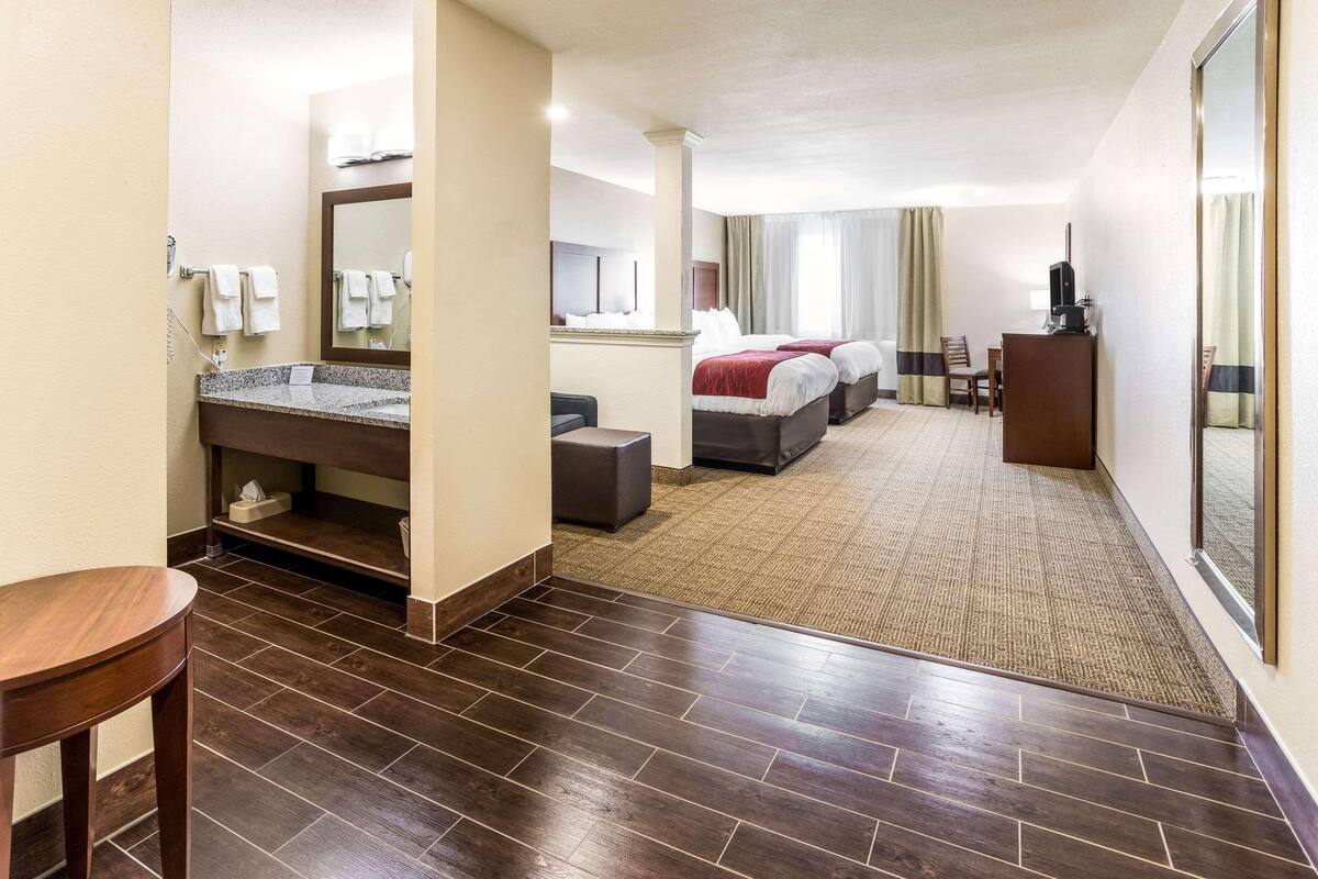 A hotel room with a large bed, a desk, a chair, a small table, a bathroom with a sink and mirror, and a carpeted floor.