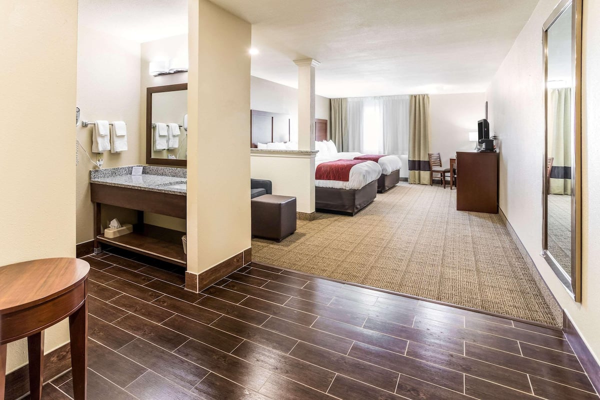 Une chambre d’hôtel avec un grand lit, un bureau, une chaise, une petite table, une salle de bain avec un lavabo et un miroir, et un sol recouvert de moquette.