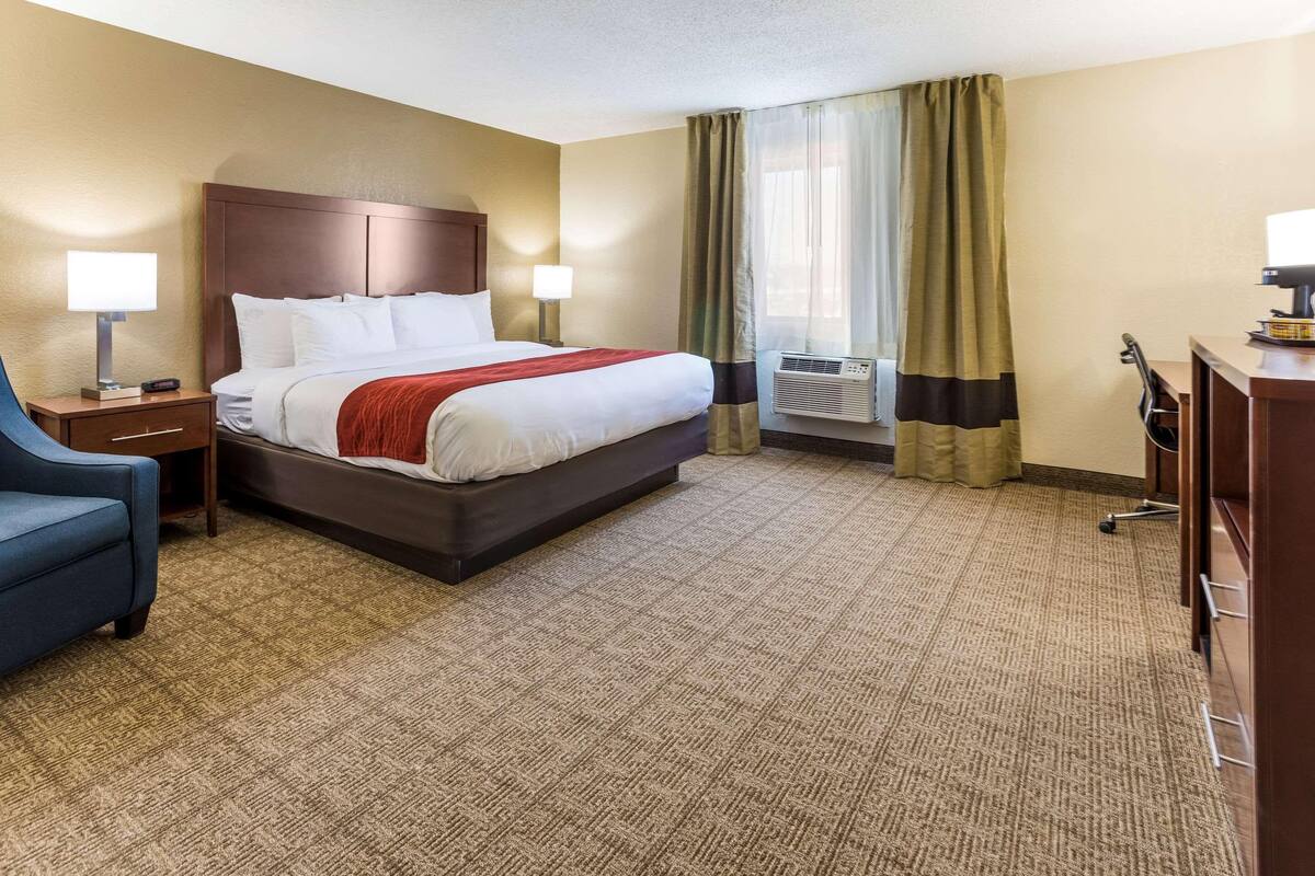 A hotel room with a large bed, a desk, a chair, and a desk lamp.