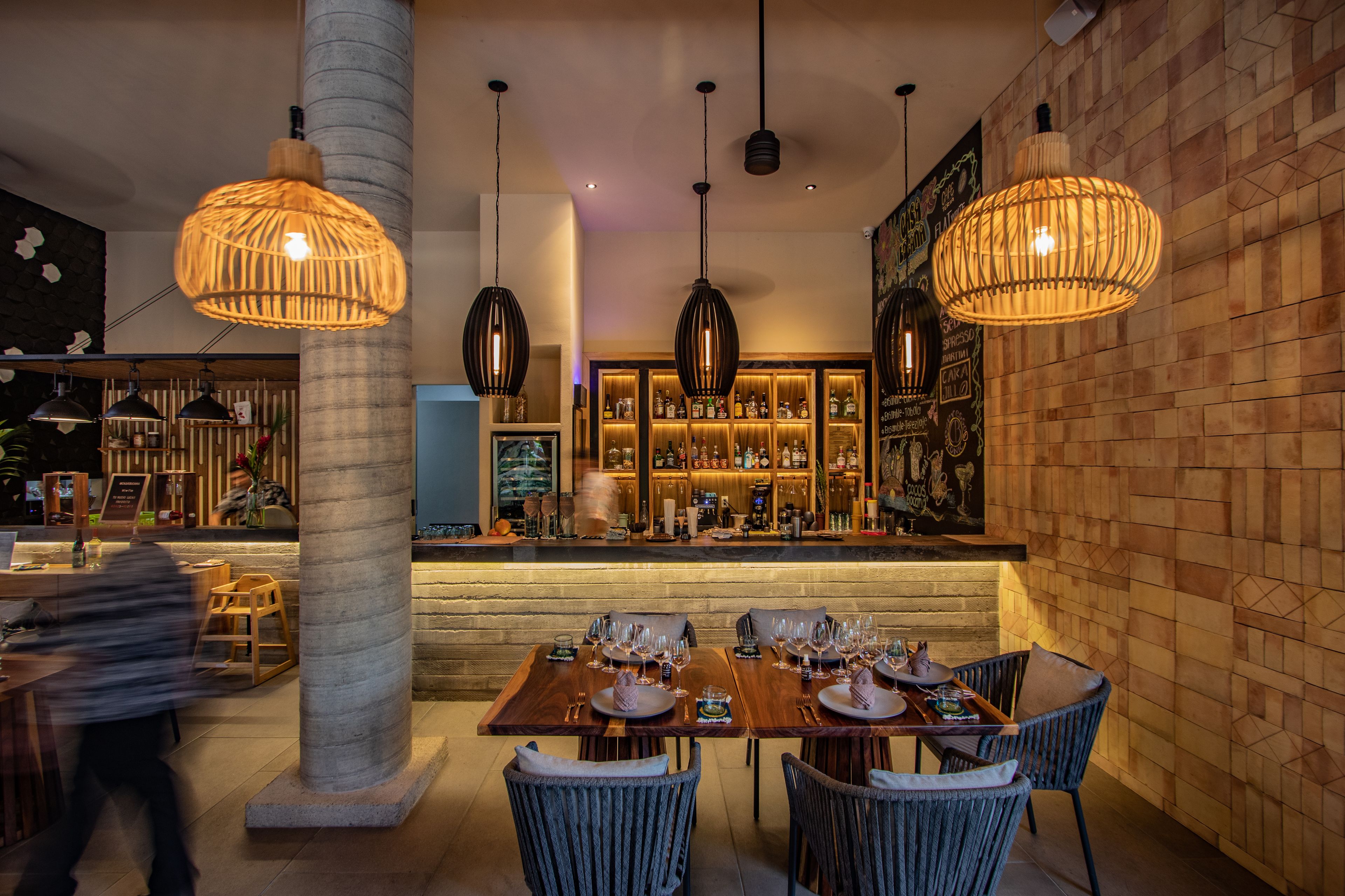 A restaurant with a bar area, wooden tables, and hanging pendant lights.
