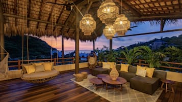 Una terraza de madera con un columpio colgante, asientos acolchados y un amplio ventanal con vistas a un paisaje montañoso.