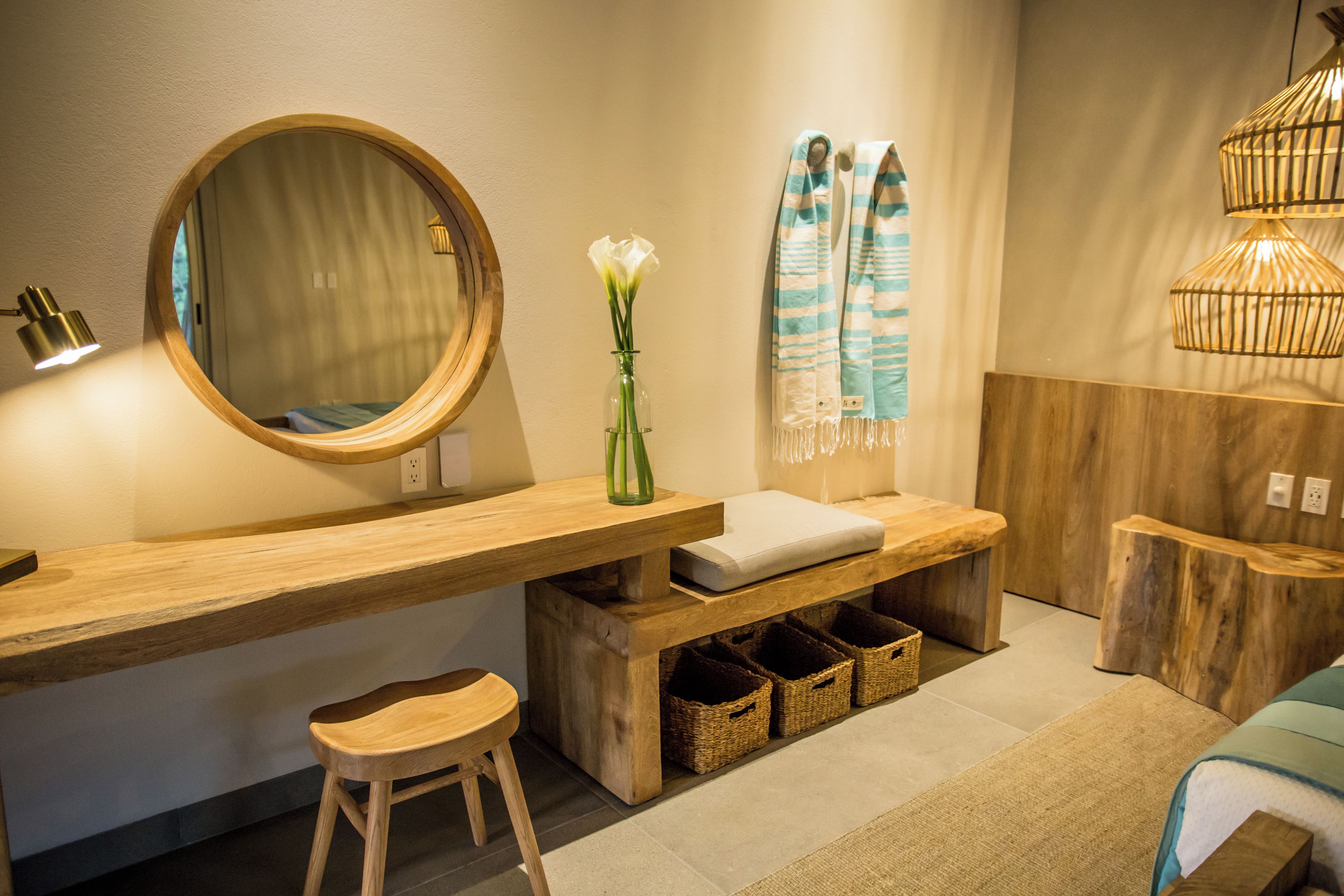 Un baño moderno con tocador de madera, un espejo redondo, un florero con flores, un perchero para toallas y una lámpara colgante.