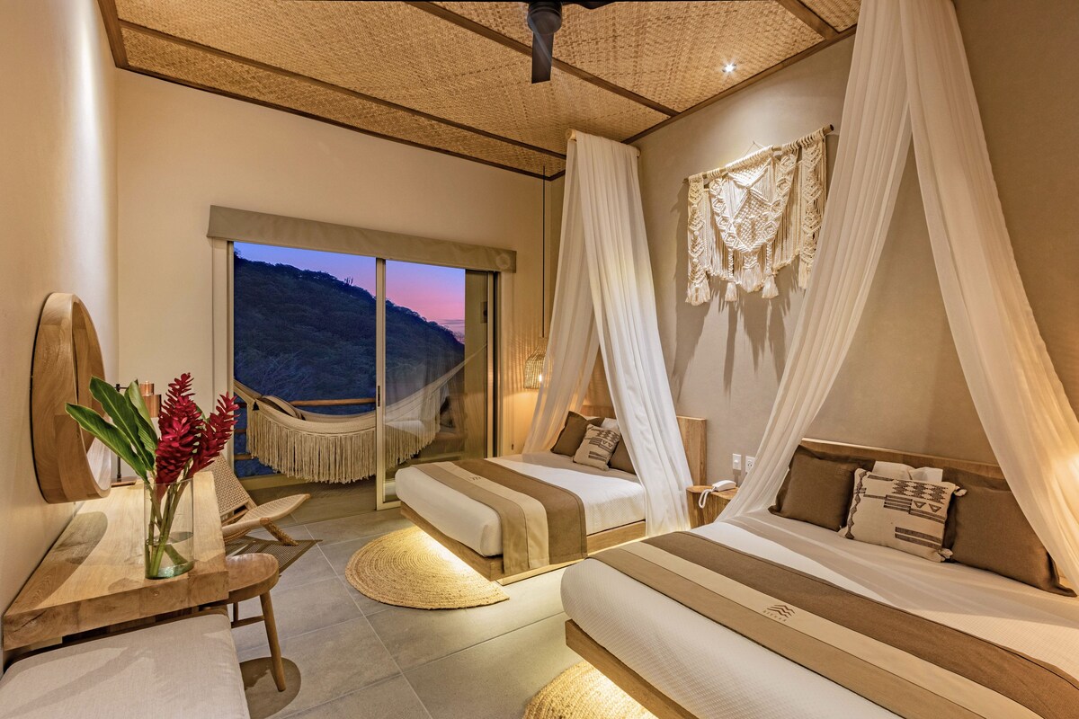 A bedroom with two beds, a large window, a hammock, and a view of the hills.