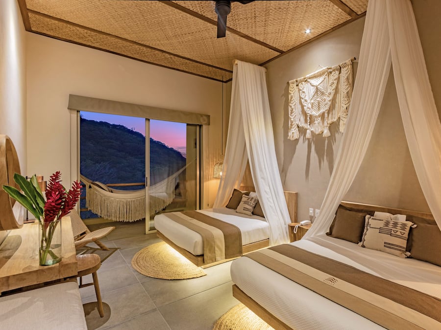 A bedroom with two beds, a large window, a hammock, and a view of the hills.