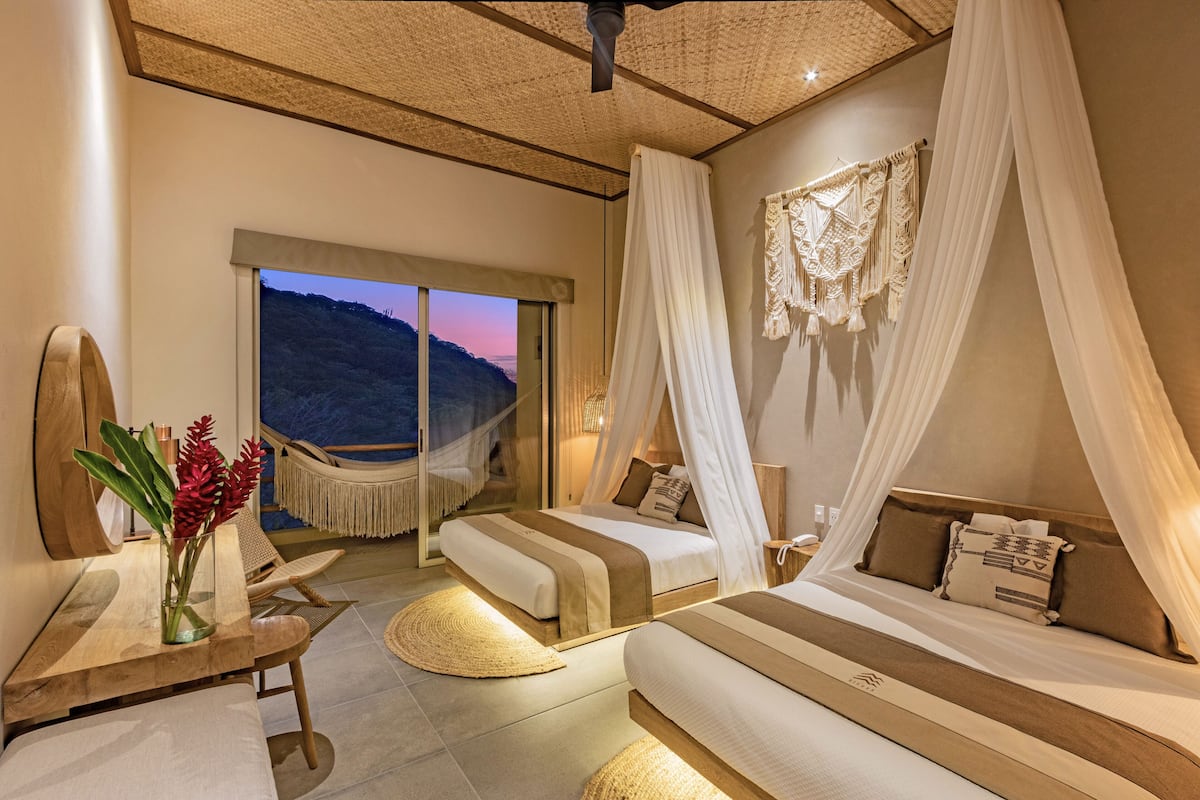 A bedroom with two beds, a large window, a hammock, and a view of the mountains.