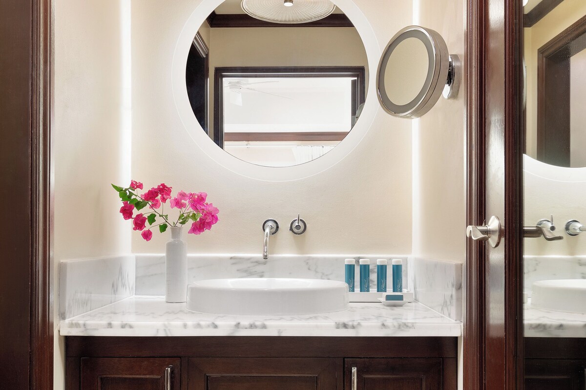Une salle de bain avec un lavabo blanc, un miroir, un vase avec des fleurs roses et des articles de toilette posés sur le comptoir.