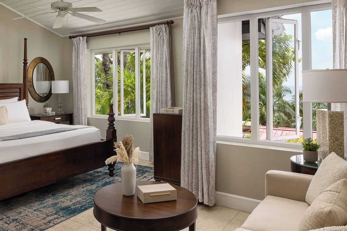 A hotel room with a large bed, a round wooden table, a nightstand, a mirror, and a view of the outdoors.
