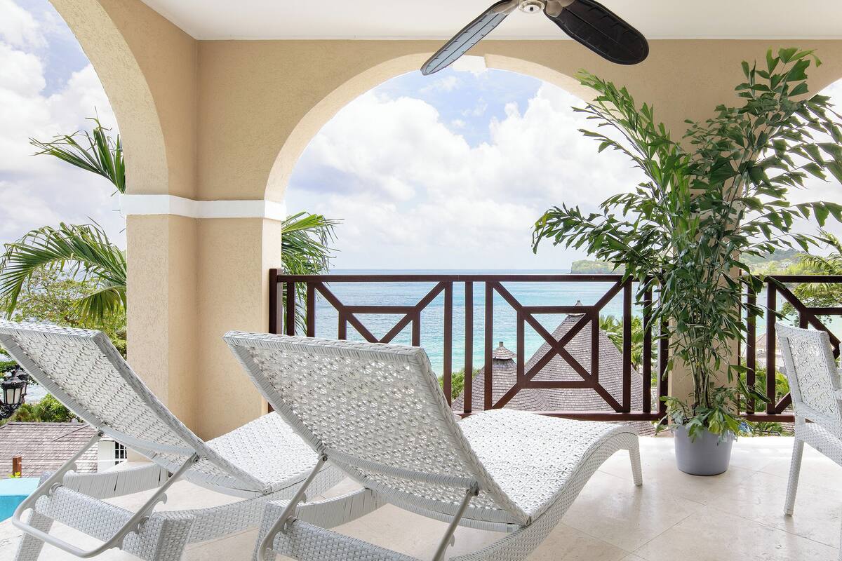 A balcony with white wicker chairs and a potted plant, overlooking a scenic view.
