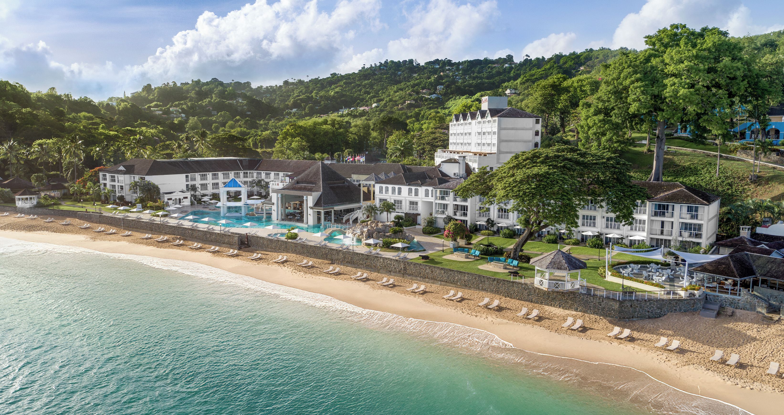 Un hotel frente al mar con piscina, playa y exuberante vegetación.